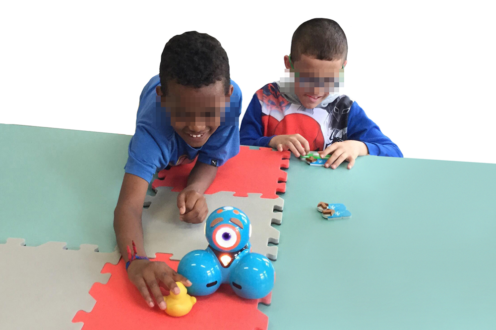 Photo taken from the Workshop with children. Two visually impaired children perform a goal-directed programming activity using a map located on the table. The activity consists of moving the robot to reach the duck. The map is made of 4 EVA foam tiles with two different colors. The child on the right is concentrated in arranging the blocks while the children at the left are touching the duck after the robot reached the duck. This child is smiling and has much of his body on the table.
