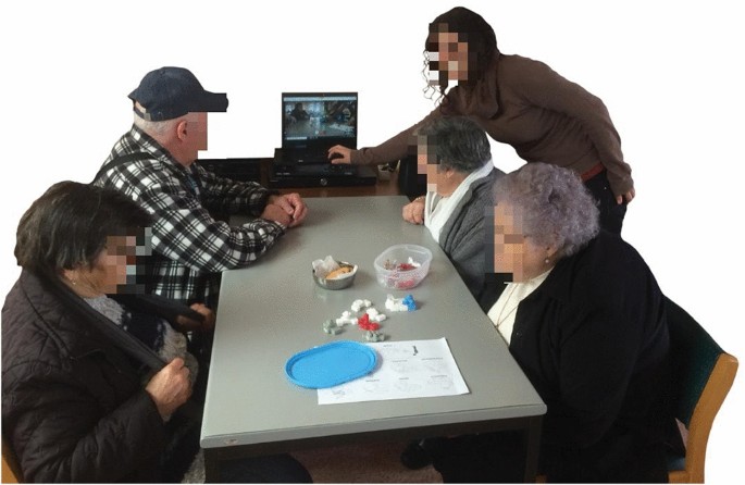 4 older adults around a table looking at the screen and listening to the researcher explaining the role of the tangible pieces on the table