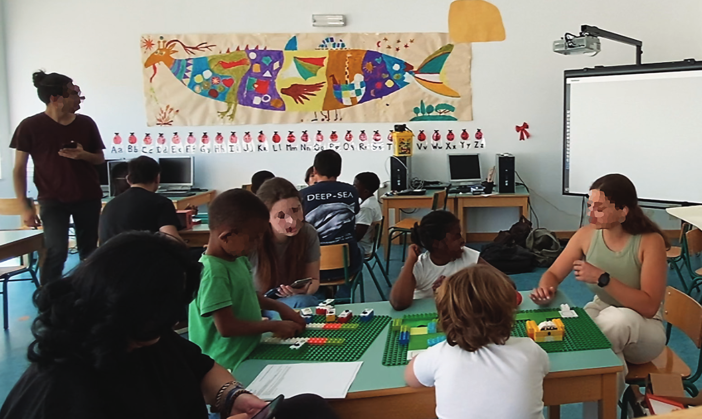 A classroom with two groups of teachers and students around one table. There is a table at the back with 4 students (1st to 4th grade) and two male teachers. A reseacher on the left of the picture moves from the back table to the front table. At the front table are 3 students (1st to 4th grade), two researchers working with them, and a teacher. On the closer table, there are LEGO-baseplates, one with a LEGO-based map and another where children are placing tangible blocks to program the robot on the map.